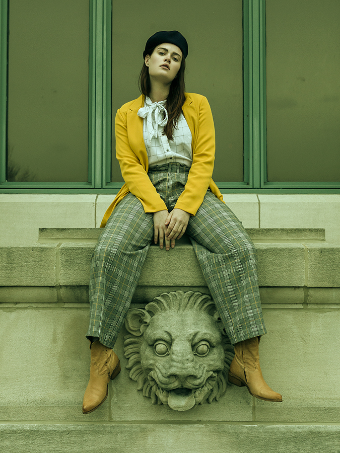Model Malia Greiner sitting on a ledge above a stone lion sculpture facade wearing plaid pants, checked blouse, yellow jacket, and beret photographed on location by Joshua Pyles